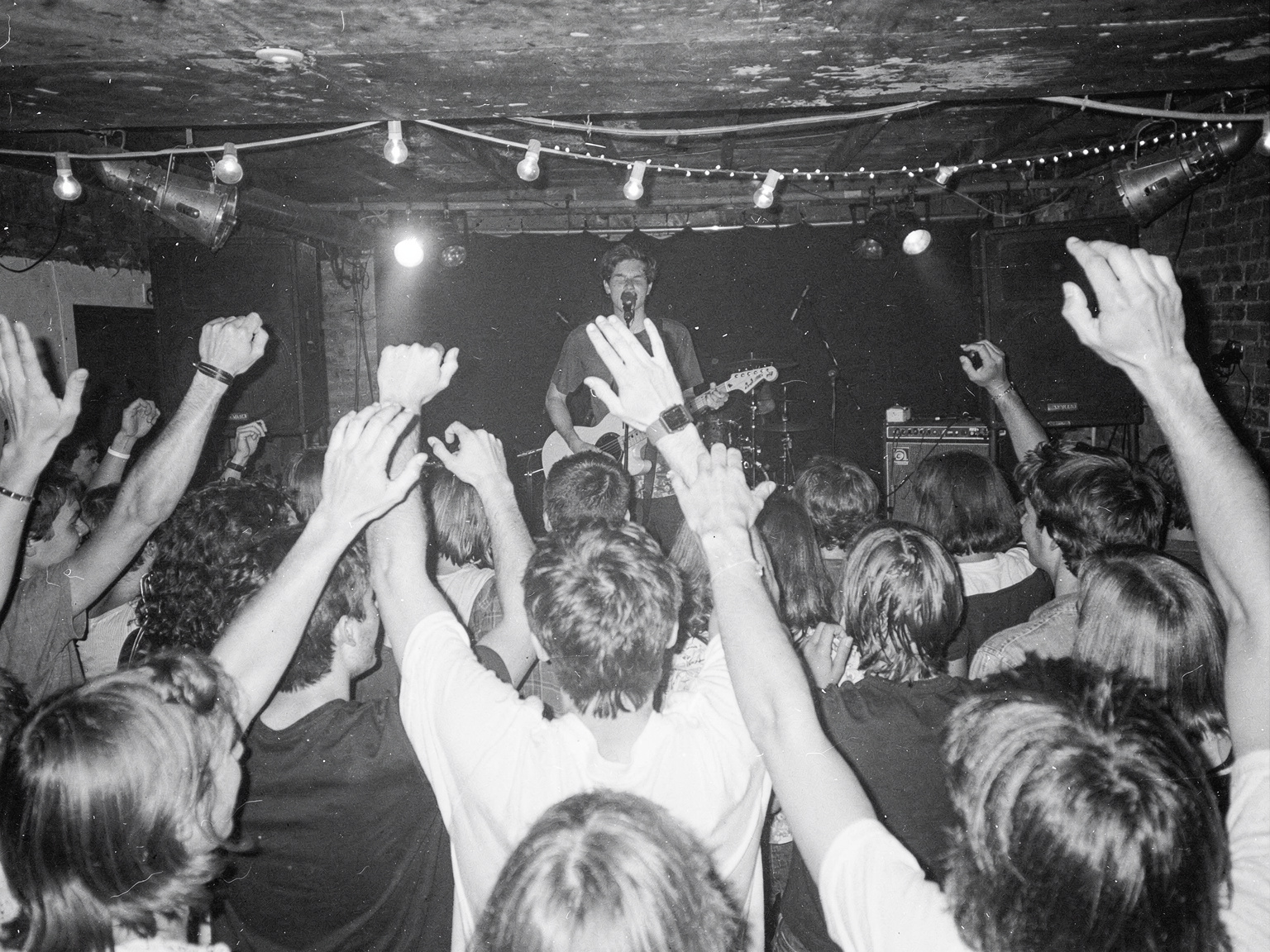Black-and-white flash photo: a crowd of teenagers with raised arms at a packed basement show, a singer with a guitar at the back under string lights.