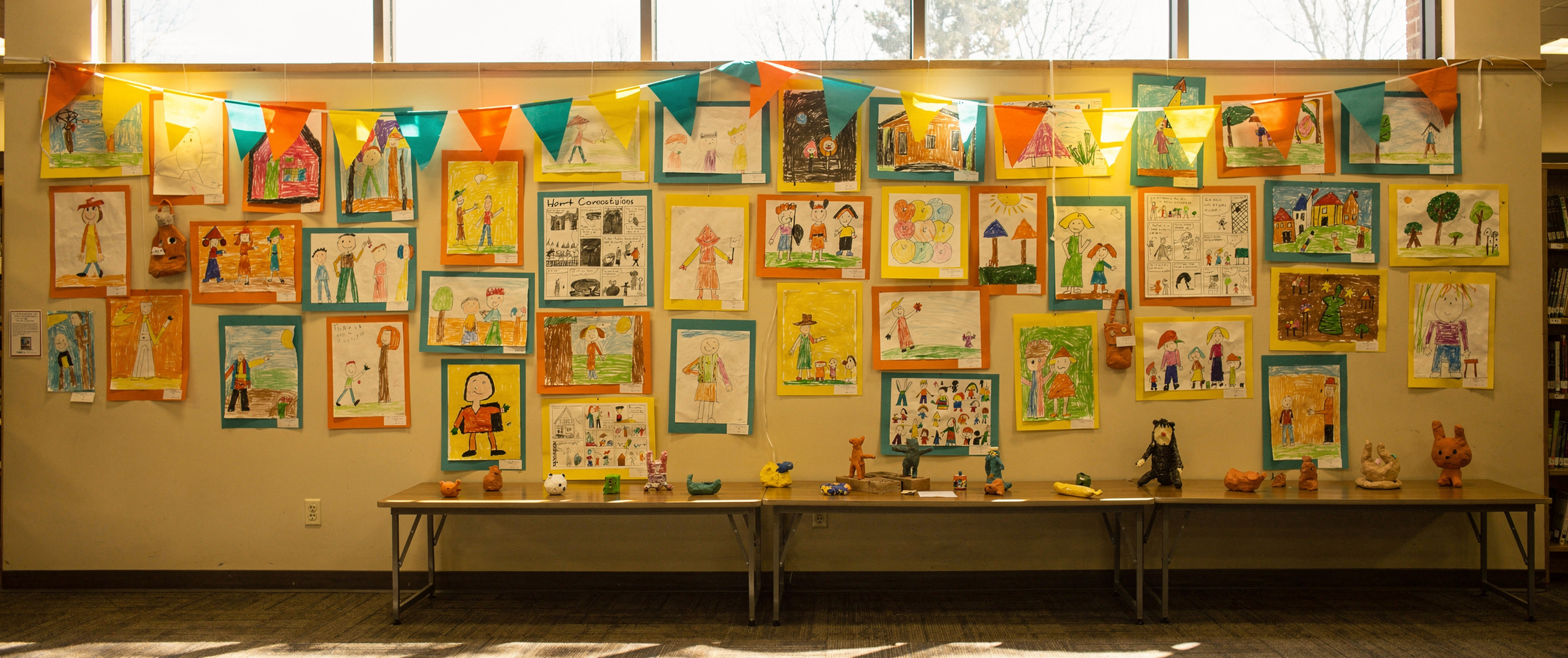 A library community room wall hung with dozens of colorful children's drawings, paintings and comics on bright mats, under orange, teal and yellow paper bunting, with small clay animals on a table below.