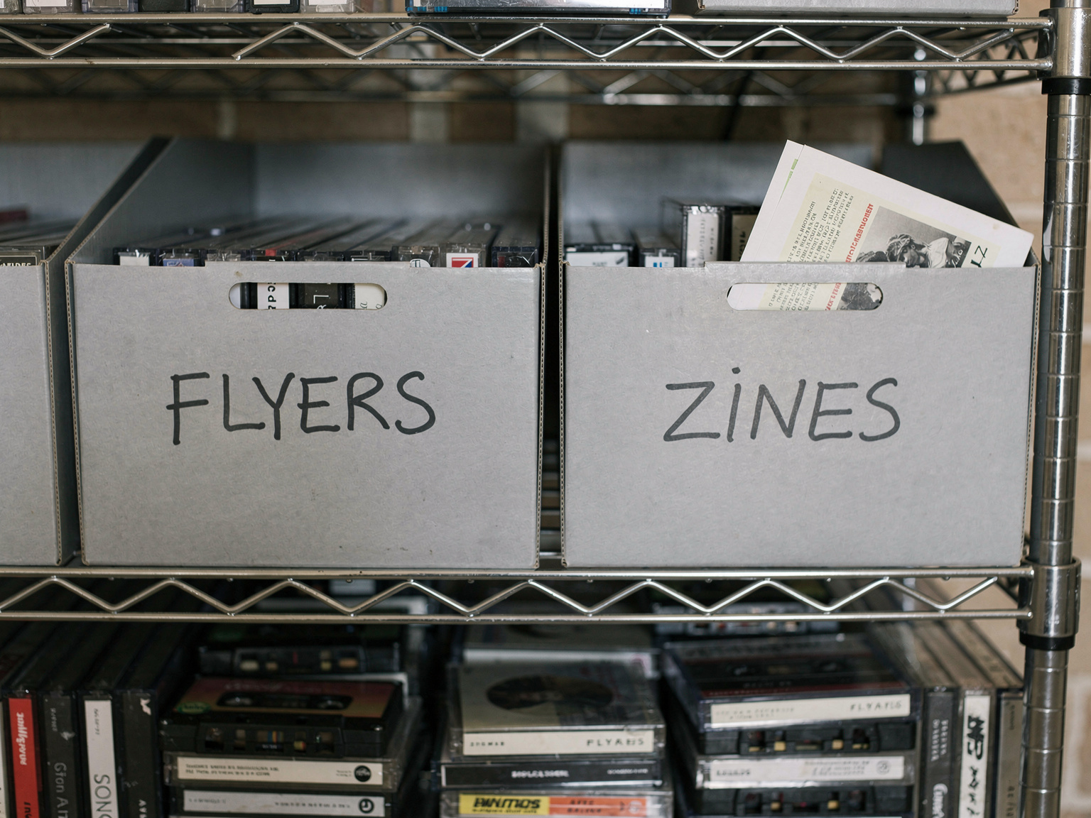 Color photo: gray archive boxes labeled flyers and zines on metal library shelves above rows of cassette tapes.