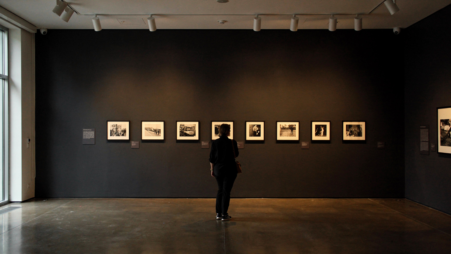 A museum gallery with dark charcoal walls and a row of small framed black and white photographs hung at eye level, warm track lighting, one visitor seen from behind looking at the photographs.