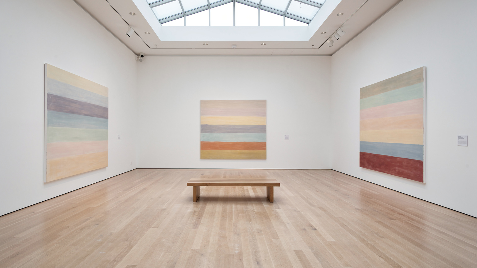 A wide view of a quiet museum gallery with white walls and a pale oak floor; three large color field paintings in soft horizontal bands hang on the walls with generous space between them; a low wooden bench in the center; daylight from a clerestory window.