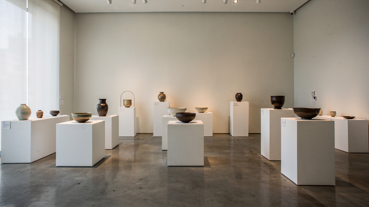 A museum gallery with ceramic vessels and bowls on white plinths of different heights, soft spotlights, pale walls and a polished concrete floor.