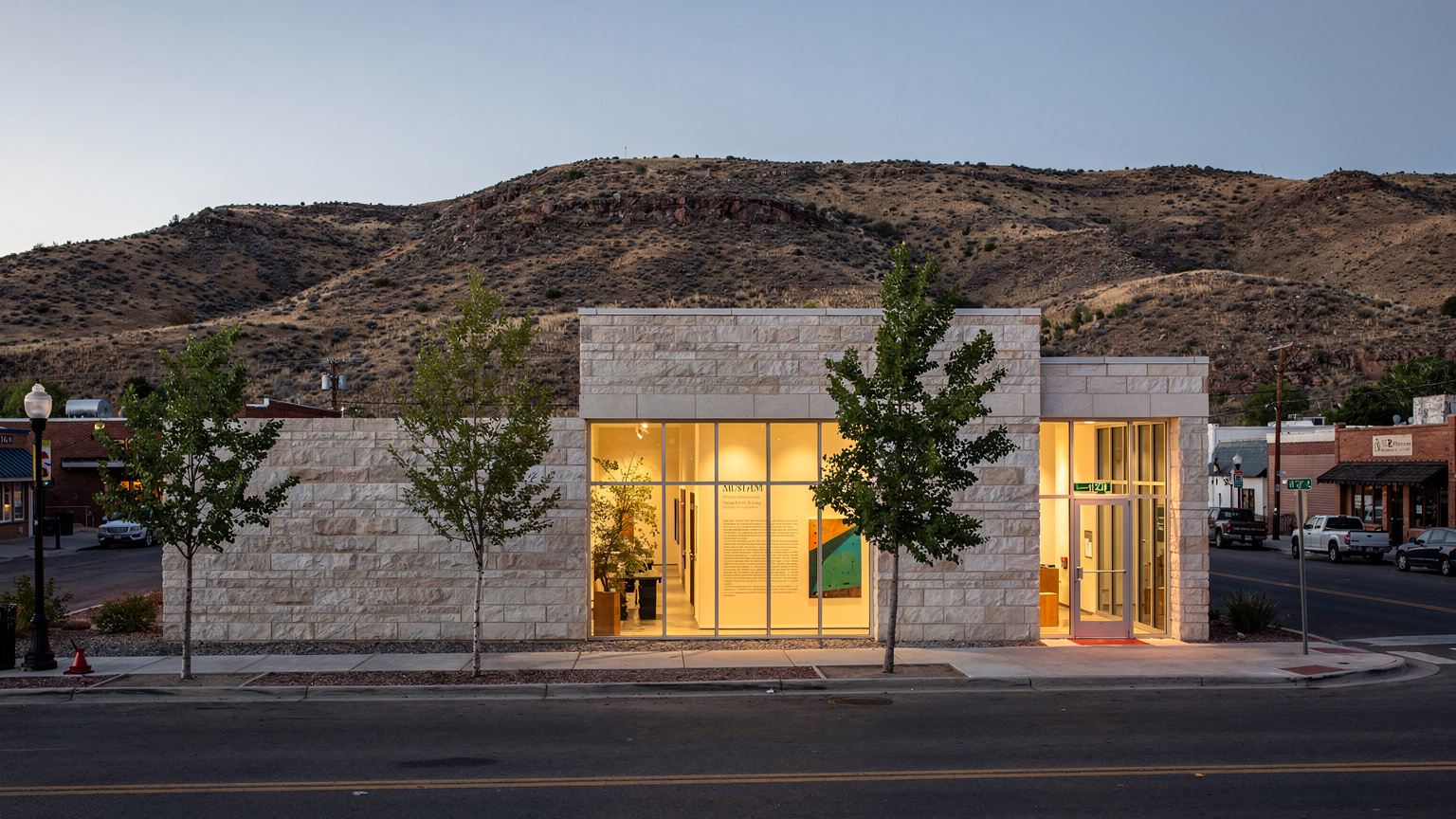 A small modern art museum building of pale stone and glass at dusk in a small western American town, warm light glowing inside, dry hills behind, a few young trees in front.