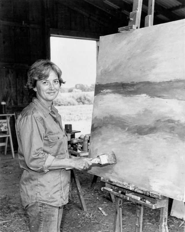 Mara Lind in her studio, about 1975. A black and white photograph from the mid 1970s of a woman painter in her late forties with short grey-streaked hair and a work shirt, standing in a barn studio on a farm beside a large unfinished canvas of soft horizontal bands of color, holding a wide brush, daylight from a big open barn door.