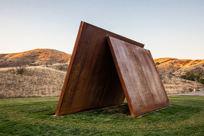 A large outdoor abstract sculpture of two rusted weathering steel plates leaning against each other on a mown lawn, dry golden hills behind, clear evening light.