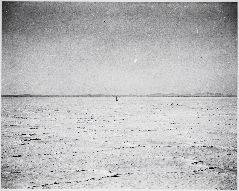 A black and white gelatin silver photograph from the 1970s of a vast flat salt flat under a pale empty sky, a thin horizon line and one tiny distant standing figure, fine film grain.