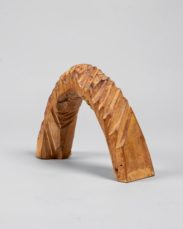 An abstract carved cedar wood sculpture shaped like a leaning arch, visible chisel marks and warm wood grain.