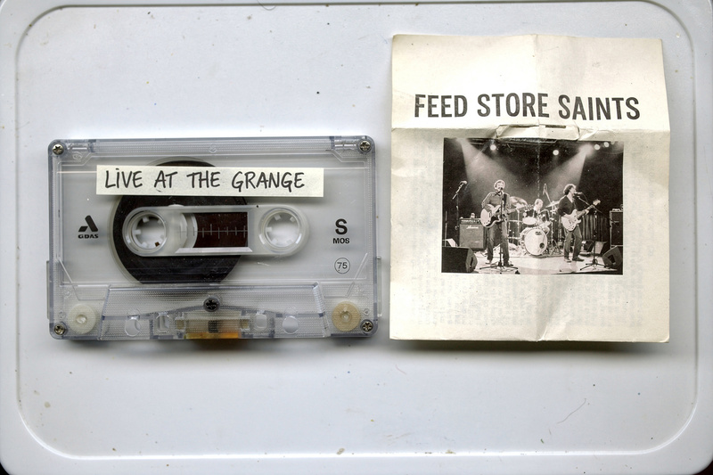 A clear cassette labeled Live at the Grange beside its J-card with a photocopied photo of a band and the words Feed Store Saints.