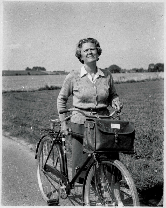 A 1950s black and white portrait photograph of a middle-aged woman in a cardigan standing beside a bicycle with a mail bag on a country road, squinting into the sun, fields behind her, film grain.