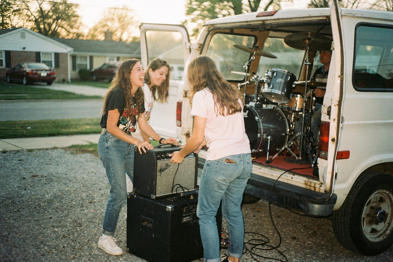 Color photo: three young women load amplifiers and a drum kit into an old white van in a gravel driveway in early morning light.