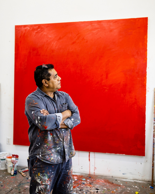 Rafael Ortiz-Bell in his studio, 2006. A color photograph of a Latino man in his forties in paint-spattered work clothes, standing with arms folded in a large white-walled studio in front of an enormous cadmium red canvas, looking at the painting.
