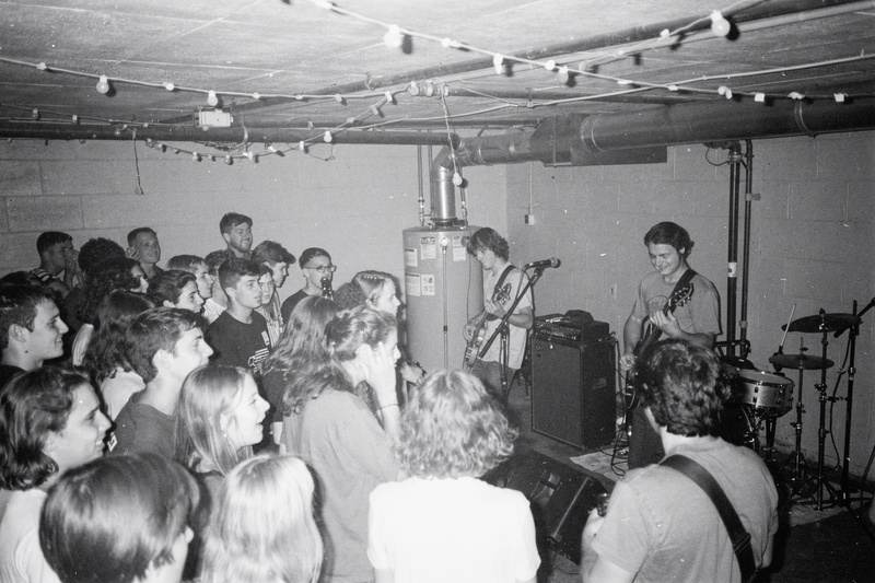 Black-and-white flash photo: teenagers pack a low basement, singing along to a band playing in the corner next to a water heater.