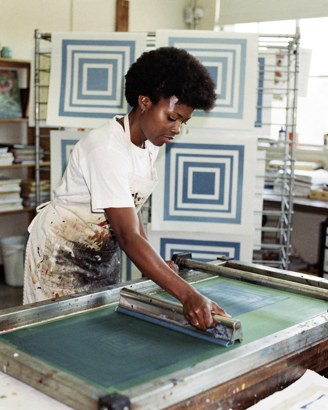 Ines Okafor in the print studio at Wrenfield College, 1991. A color photograph from the early 1990s of a Black woman printmaker in her thirties with short natural hair and an ink-stained apron, pulling a squeegee across a silk screen in a bright print studio, freshly printed sheets of nested blue squares drying on a rack behind her.