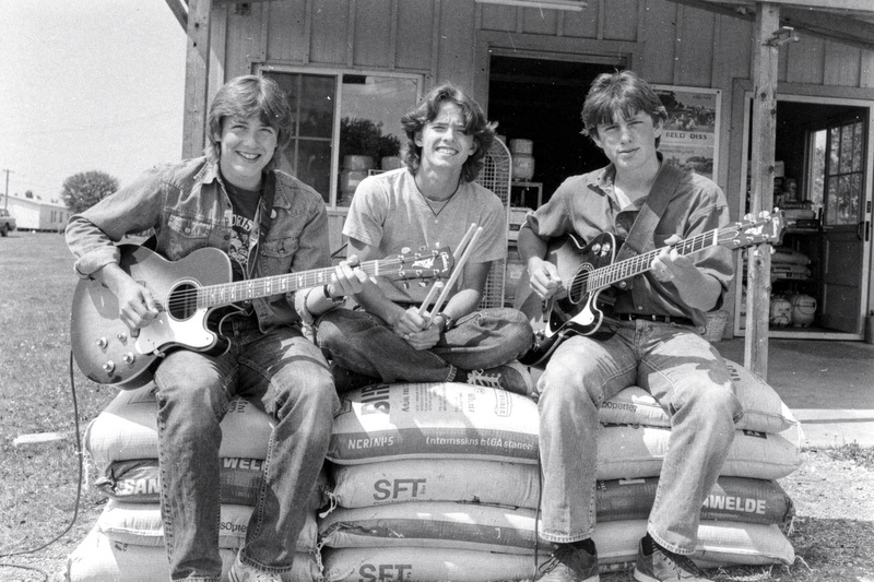 Black-and-white photo: three teenage musicians sit on stacked feed sacks in front of a rural feed store, two with guitars and one with drumsticks.