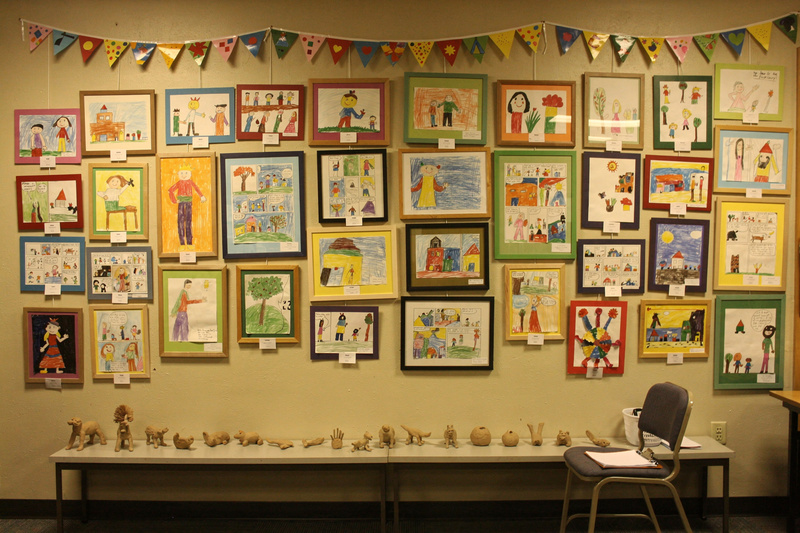 Library wall hung salon-style with dozens of children's drawings, paintings, collages and comics, paper bunting above and small clay animals on a shelf below.