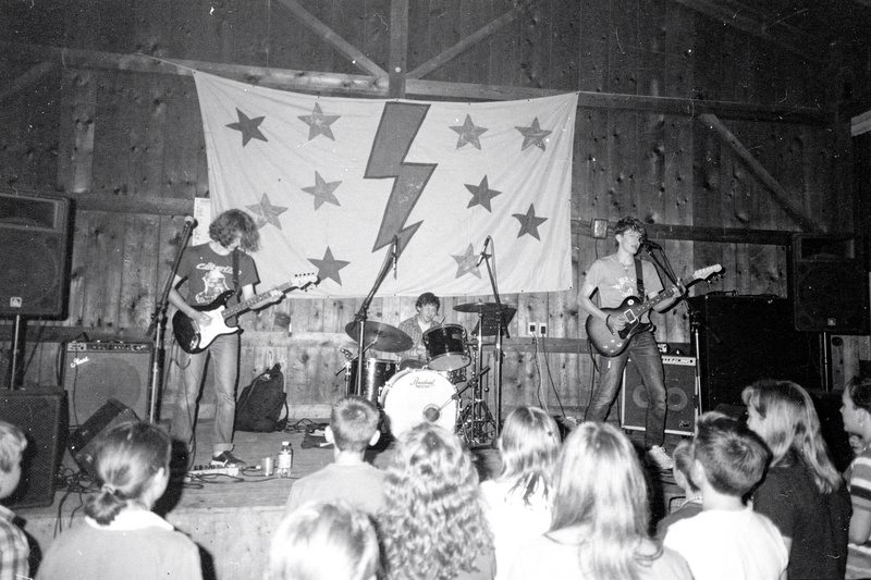 Black-and-white flash photo: a teenage band plays on a small wooden stage in a grange hall under a banner painted with stars and a lightning bolt, kids crowded at the front.