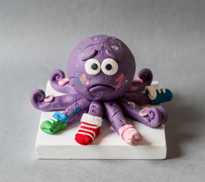 Purple clay octopus with worried eyebrows; six arms wear painted clay socks, one wears a real striped baby sock, and one arm is bare.