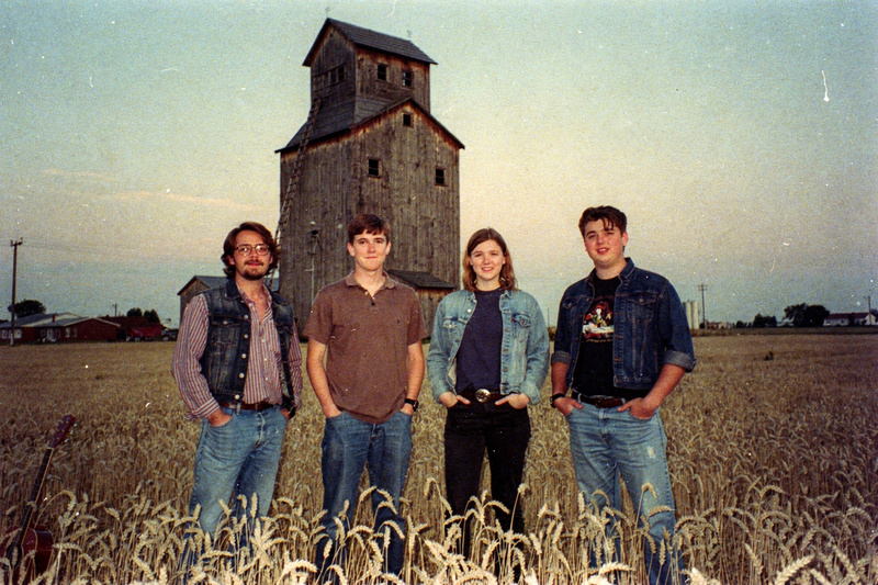 Faded color photo: four young musicians stand in a wheat field at dusk with a tall grain elevator behind them.