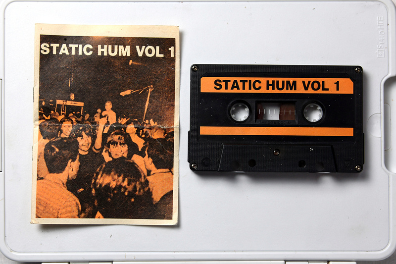 A black cassette labeled Static Hum Vol. 1 beside its J-card, an orange-and-black halftone photo of a crowd at a show.
