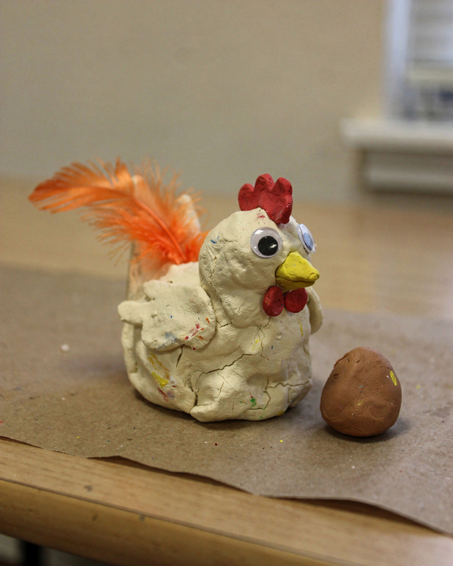 Very lumpy cream clay chicken with two different-size googly eyes, a red comb, orange craft feathers and a small brown clay egg beside her.