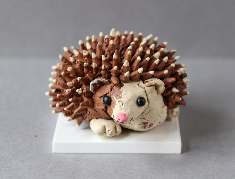 Clay hedgehog curled up a little, its back covered in hundreds of snipped clay spikes painted brown with pale tips, a cream face with bead eyes and a tiny pink nose.