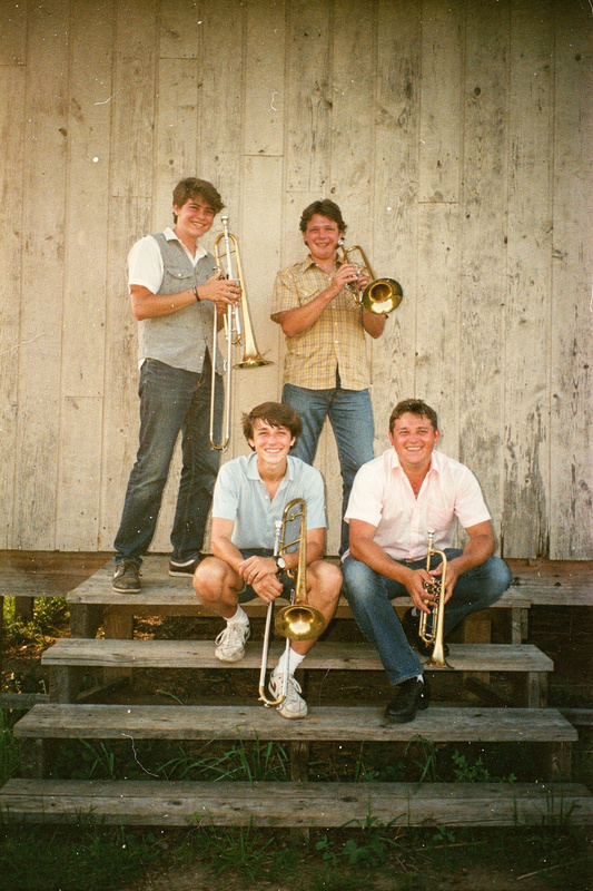 Color photo: four young musicians grin on the wooden steps of the grange hall, holding two trombones and two trumpets.