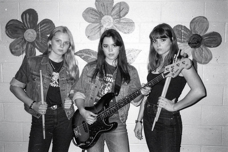 Black-and-white flash photo: three young women pose against a cinderblock wall painted with big flowers, one holding a bass guitar and one holding drumsticks.