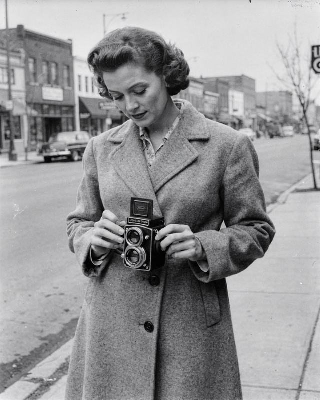 Ruth Calder on Main Street, 1955. A black and white photograph from the 1950s of a woman in her thirties with short wavy hair and a wool coat, holding a twin-lens reflex camera at waist height on the sidewalk of a small-town main street, looking down into the camera's viewfinder.