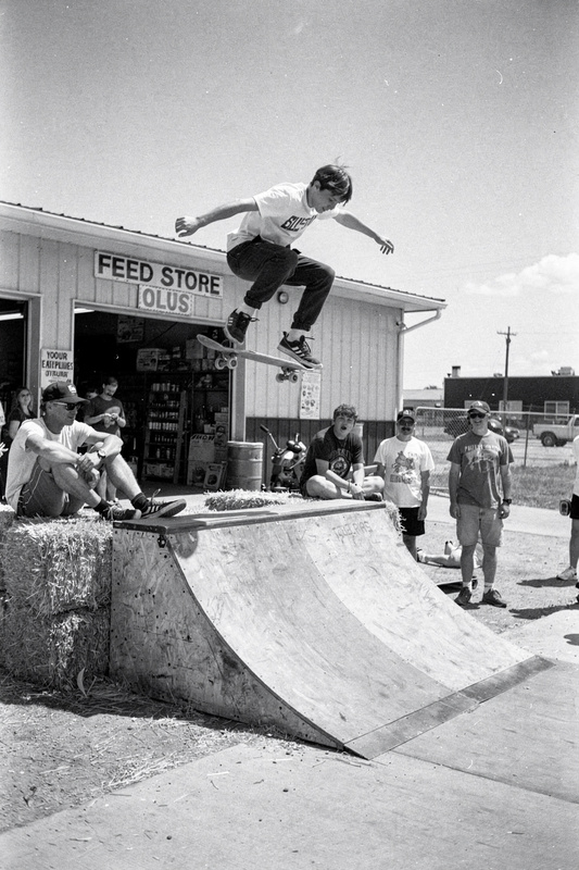 Black-and-white photo: a teenage skateboarder flies above a homemade plywood ramp behind a feed store while friends watch from hay bales.