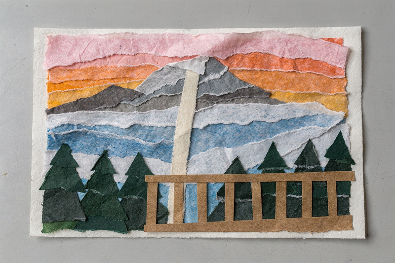Torn-paper collage of a snowy mountain at sunset seen over a porch railing, with pine forests, a pale cleared stripe down the slope, and a pink and orange tissue-paper sky.