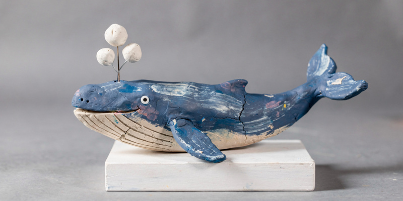 Long clay blue whale with a pale belly, scratched throat lines, a smiling eye and three white clay bubbles on a wire above its blowhole.