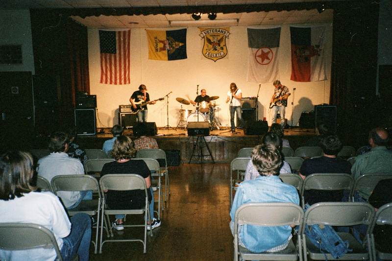 Color photo: a four-piece band plays under warm light on the stage of a veterans hall while the audience sits on rows of folding chairs.