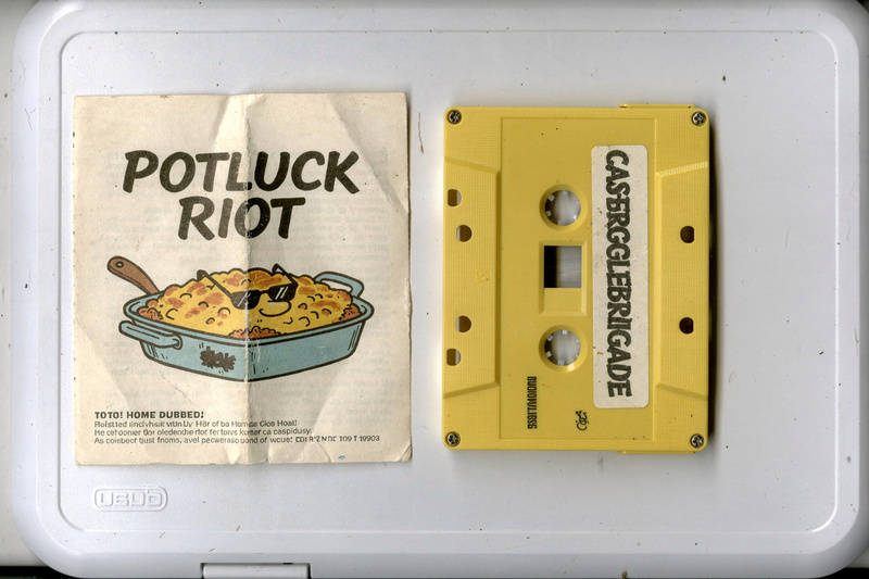 A yellow cassette labeled Casserole Brigade beside its J-card with a cartoon of a casserole dish in sunglasses and the words Potluck Riot.