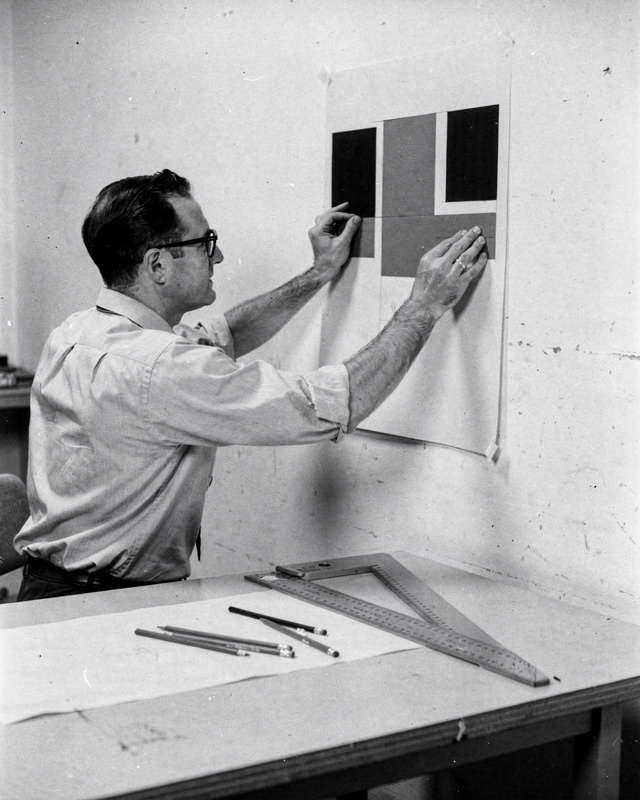 Tomas Ekwall at his drafting table, 1966. A black and white photograph from the 1960s of a man in his thirties with glasses and rolled shirt sleeves, seated at a drafting table in a sparse studio, pinning a study of black, red and grey rectangles to the wall, a T-square and pencils on the table.
