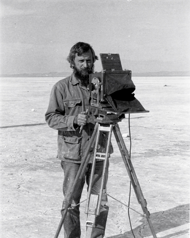 Sol Becker on the salt flats, 1976. A black and white photograph from the 1970s of a bearded man in his thirties in a canvas work jacket, standing behind a large-format wooden view camera on a tall tripod on a vast flat salt plain, holding the dark focusing cloth, a pale empty horizon behind him.