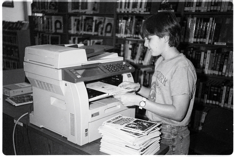 Black-and-white photo: a teenager with short hair feeds paper into a big photocopier in a library, a stack of flyers beside them.