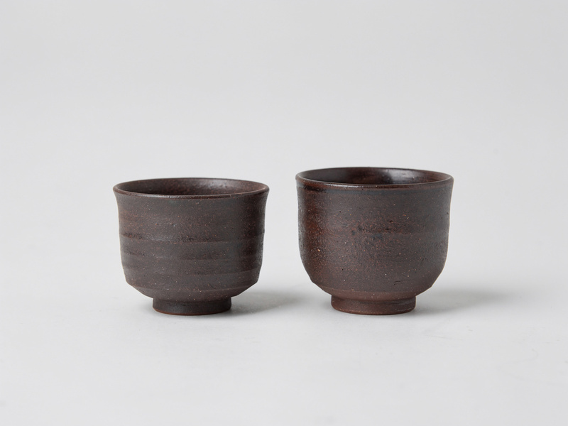 Two small handleless stoneware tea cups with a dark iron-brown glaze, one slightly taller than the other, side by side.