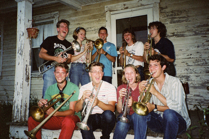 Color flash photo: about ten young people crowd onto the porch of an old wooden house with trombones and trumpets, laughing.