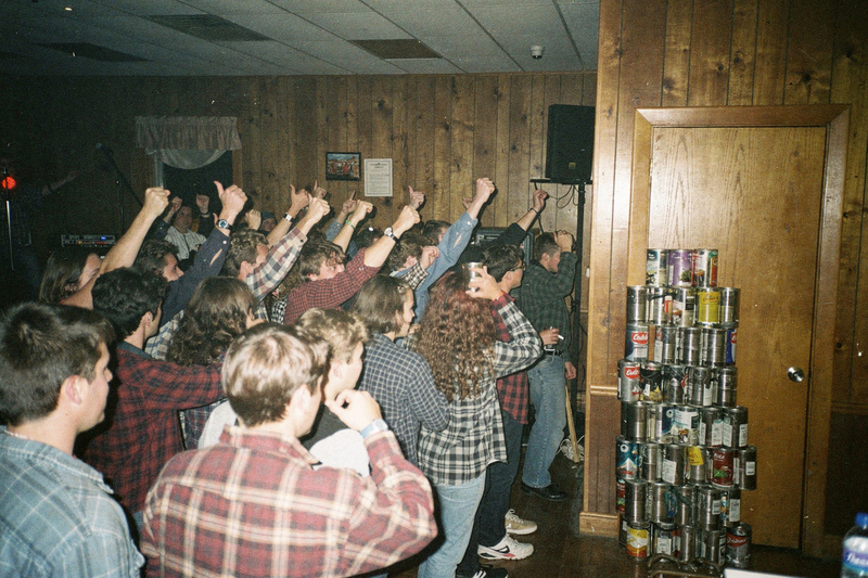 Color flash photo from the stage: teenagers in flannel shirts pack a wood-paneled lodge hall with their arms raised, tin cans stacked by the door.