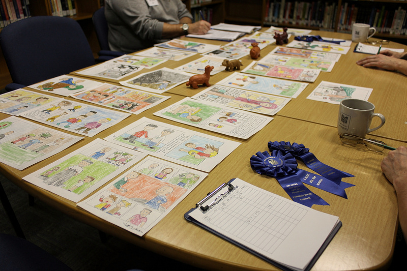 Meeting room table covered with rows of children's drawings, collages and comics, small clay animals, blue prize ribbons and a clipboard of score sheets.