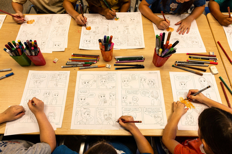 Overhead view of a library table covered in comic templates, half-finished comic pages and cups of markers, with children's hands drawing.