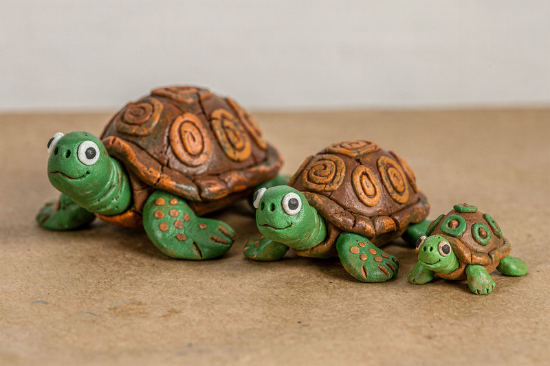 Three polymer clay turtles in a line, a mother and two babies, with green bodies and brown and orange shells patterned with toothpick dots and swirls.
