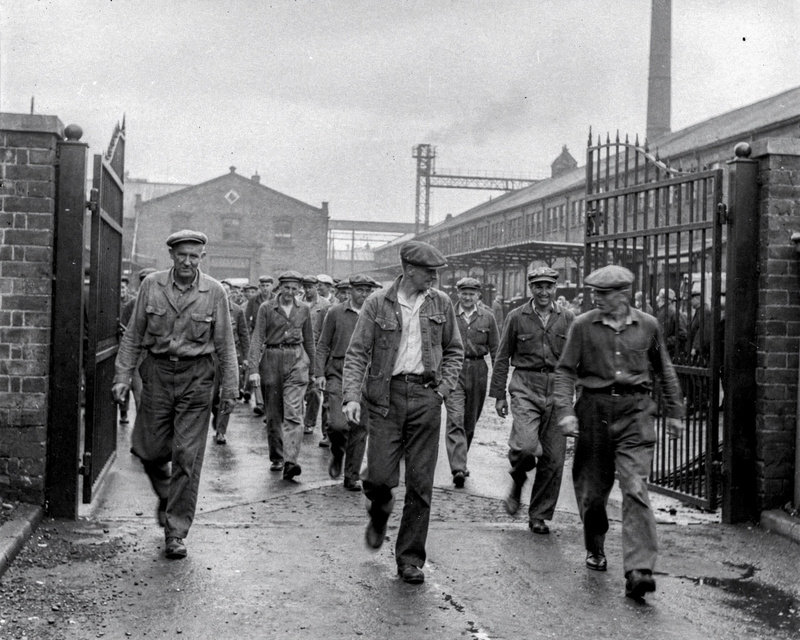 A 1950s black and white documentary photograph of mill workers in work clothes and caps walking out through a factory gate at the end of a shift, overcast light, candid, film grain.