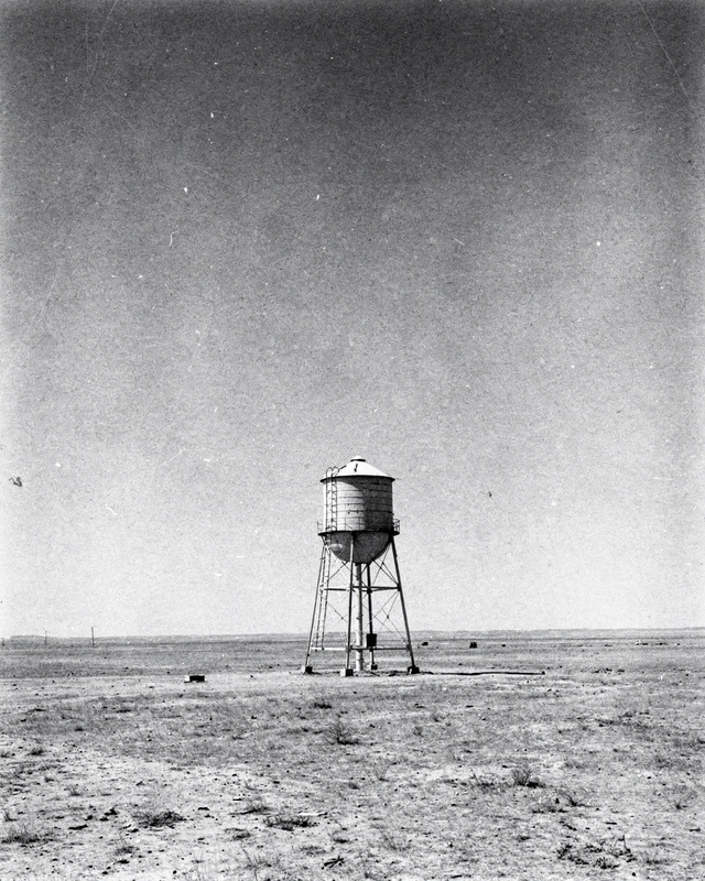 A black and white photograph of a lone steel water tower standing small on a flat empty plain under a big sky, strong midday light, fine grain.