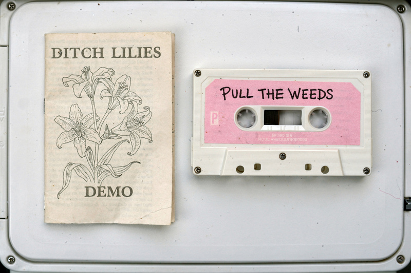 A white cassette with a pink label reading Pull the Weeds beside its J-card with a drawing of lilies and the words Ditch Lilies demo.