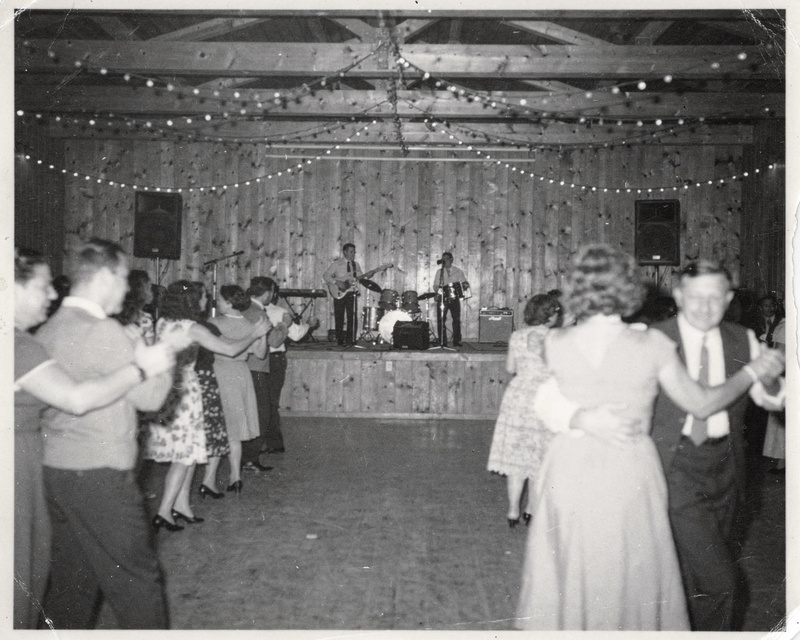 A 1960s black and white flash photograph inside a wooden community hall during a dance, couples blurred in motion, a small band on a low stage in sharp focus, strings of lights, film grain.