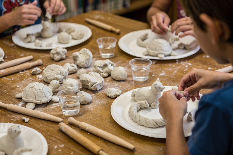 Library craft table with lumps of air-dry clay, wooden tools, water cups and paper plates of unfinished clay animals, with children's hands pinching clay.
