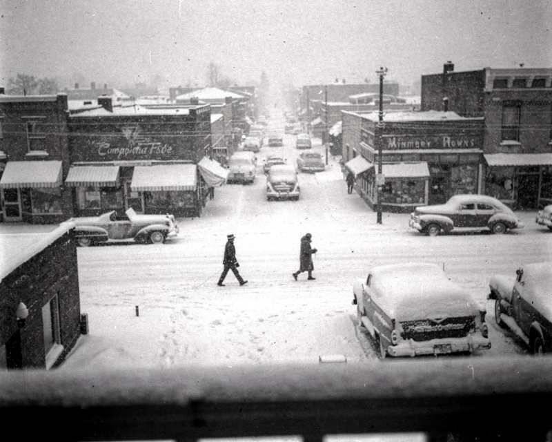 A 1950s black and white photograph taken from a second-floor window looking down a small-town main street in heavy snow, two figures crossing the street, old cars covered in snow, storefronts with awnings, film grain.