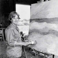 Mara Lind in her studio, about 1975. A black and white photograph from the mid 1970s of a woman painter in her late forties with short grey-streaked hair and a work shirt, standing in a barn studio on a farm beside a large unfinished canvas of soft horizontal bands of color, holding a wide brush, daylight from a big open barn door.
