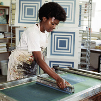 Ines Okafor in the print studio at Wrenfield College, 1991. A color photograph from the early 1990s of a Black woman printmaker in her thirties with short natural hair and an ink-stained apron, pulling a squeegee across a silk screen in a bright print studio, freshly printed sheets of nested blue squares drying on a rack behind her.